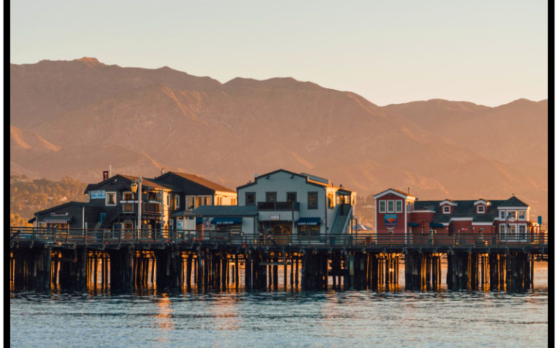 Stearns Wharf in Santa Barbara, California, at sunset with colorful waterfront buildings and coastal mountains in the background.
