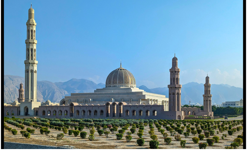 The Sultan Qaboos Grand Mosque in Muscat, Oman, showcasing its grand dome, elegant minarets, and stunning mountain backdrop under a clear sky.