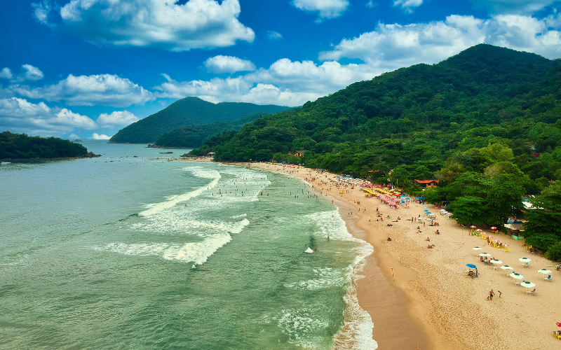 Florianópolis beach in Brazil with surfers and sunbathers enjoying warm weather, surrounded by lush green hills and turquoise waves.