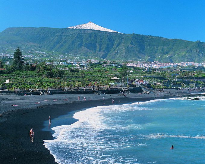 Black sand beach in Tenerife, Canary Islands, surrounded by lush green hills and coastal towns under a mild sunny sky.