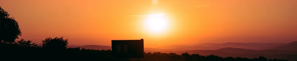 Bright orange sunset over rolling hills with a glowing sun above the horizon, silhouetted trees, and a dark signboard in the foreground.
