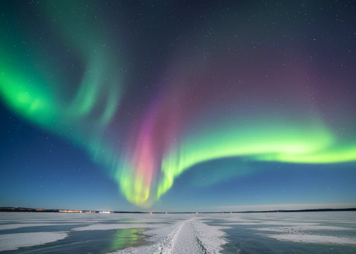 Bright green and magenta aurora reflecting on the frozen Great Slave Lake near Yellowknife, Canada, with faint city lights under a clear, starry Arctic sky.