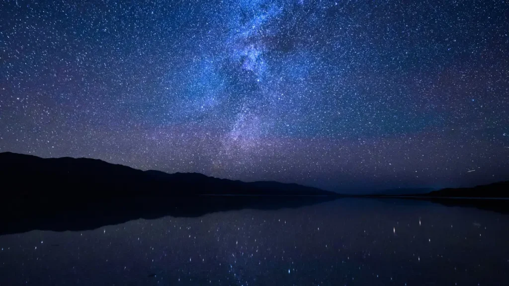 A breathtaking view of the Milky Way stretching across a clear night sky, reflected over a calm lake surrounded by distant mountain silhouettes.