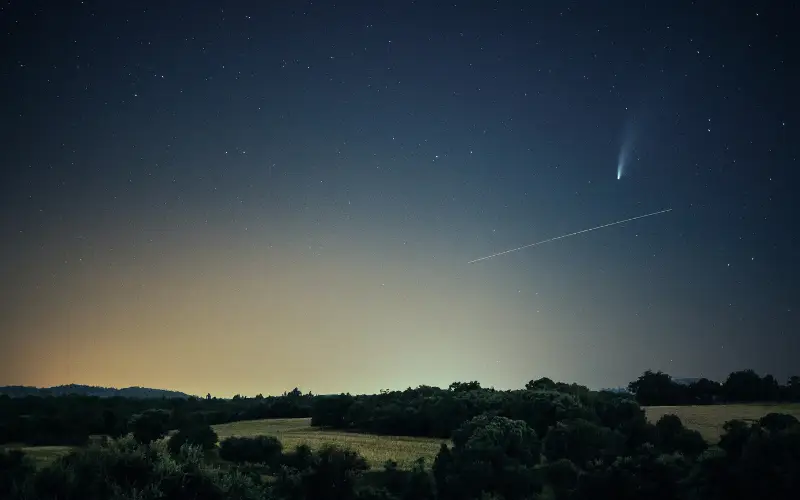 A bright meteor streaks across the clear night sky above a quiet rural landscape, with a visible comet glowing near the horizon.