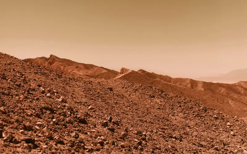 Rocky red terrain of Mars showing rugged hills and dusty plains under a hazy orange sky.