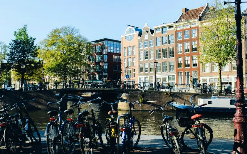 Bicycles parked along a canal in Amsterdam, Netherlands, with classic Dutch architecture and green trees, reflecting the city’s cycling culture and sustainable urban lifestyle.
