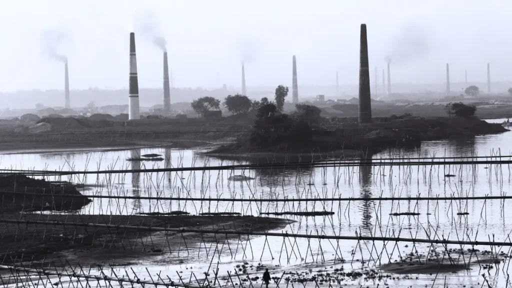Industrial smokestacks releasing pollution into the air above a river landscape, showing environmental impact and rising emissions in the region.