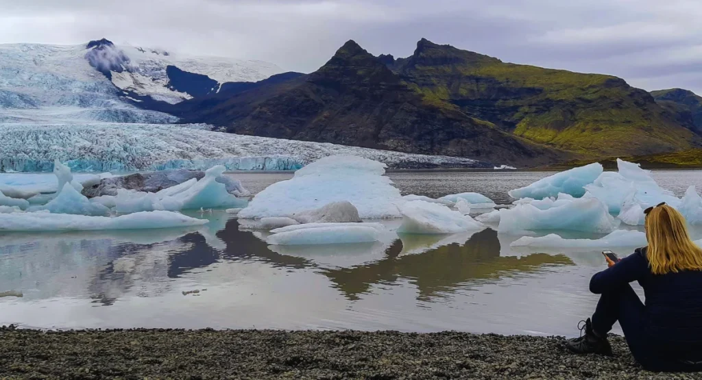 A traveler sits beside a melting glacier surrounded by floating ice in a mountain valley, symbolizing the visible effects of climate change and disappearing destinations.