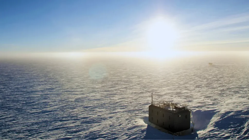 A bright sunrise over the icy expanse of Antarctica, with a remote research station sitting on the snow-covered landscape under clear blue skies.