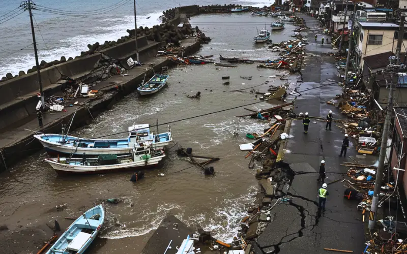 Coastal town damaged after a powerful earthquake in Japan, showing cracked roads, broken seawalls, stranded fishing boats, flooding, scattered debris, and emergency workers assessing destruction along the shoreline.